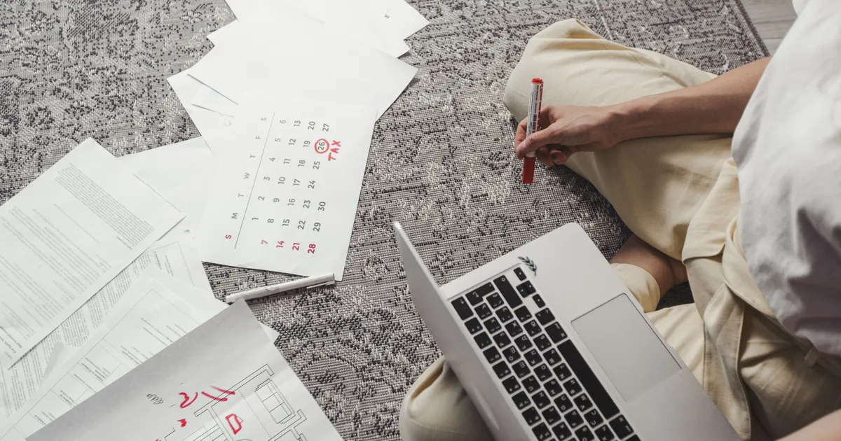 Persona con una laptop junto a un calendario y hojas de trámites extendidas sobre una alfombra