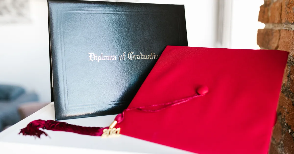 Carpeta negra de diploma de graduación junto a un birrete rojo con borla sobre una repisa blanca